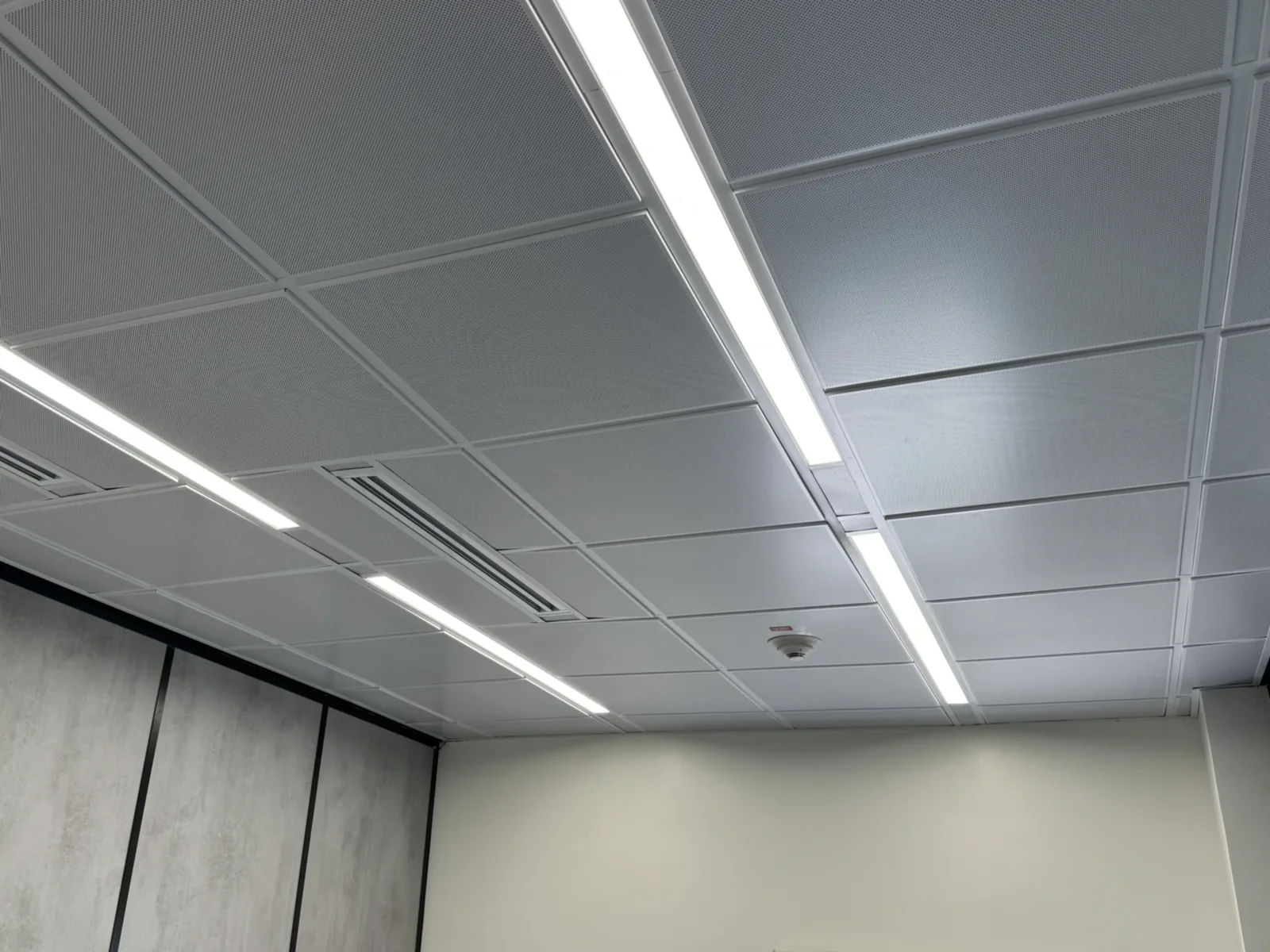 Perforated metal ceiling tiles on an exposed grid, with linear lights, slot diffusers and a smoke detector