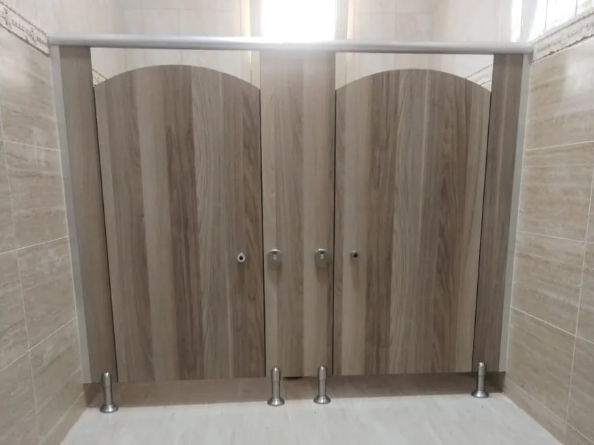 Two wood-grain toilet cubicles with arched tops under a headrail, on metal legs in a tiled washroom