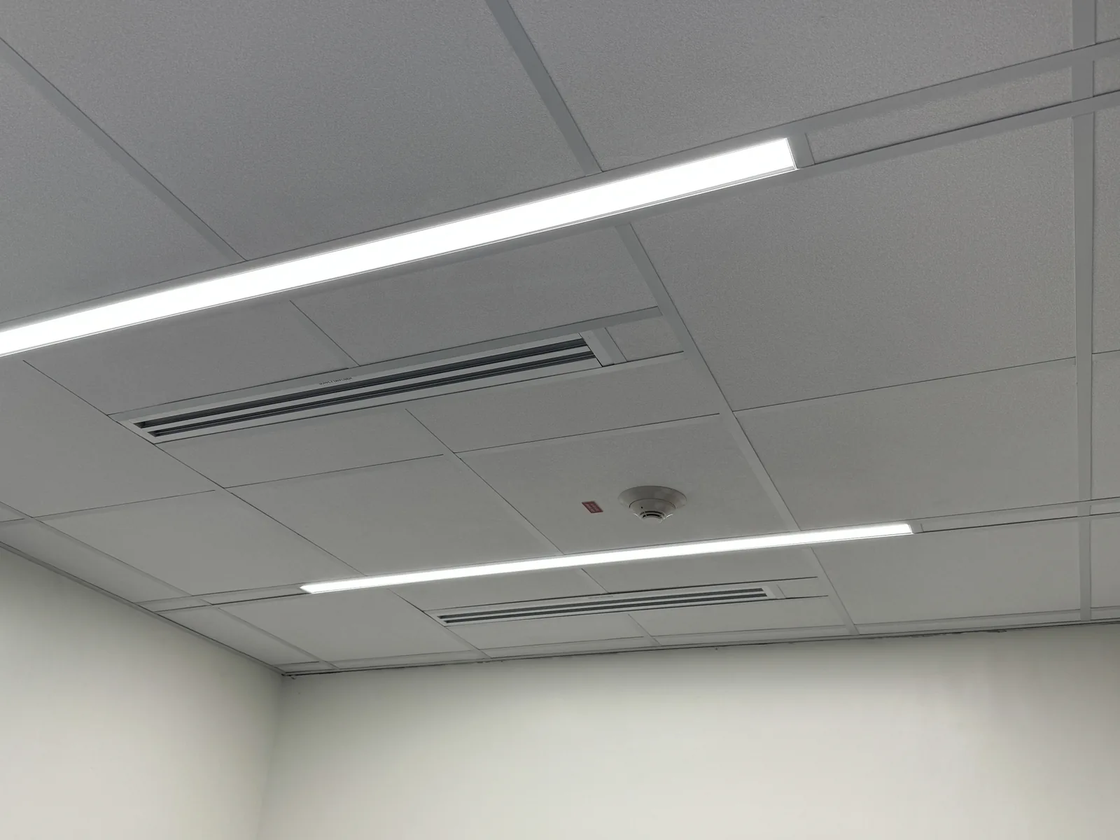 Lay-in ceiling tiles with recessed linear lights, slot diffusers and a smoke detector above white walls