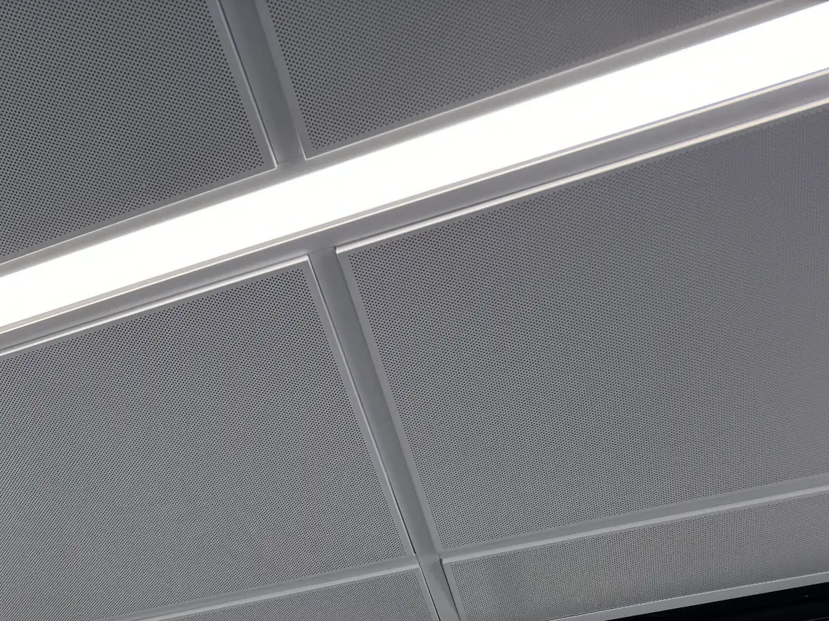 Close view of perforated metal ceiling tiles on a light grid beside a recessed linear light