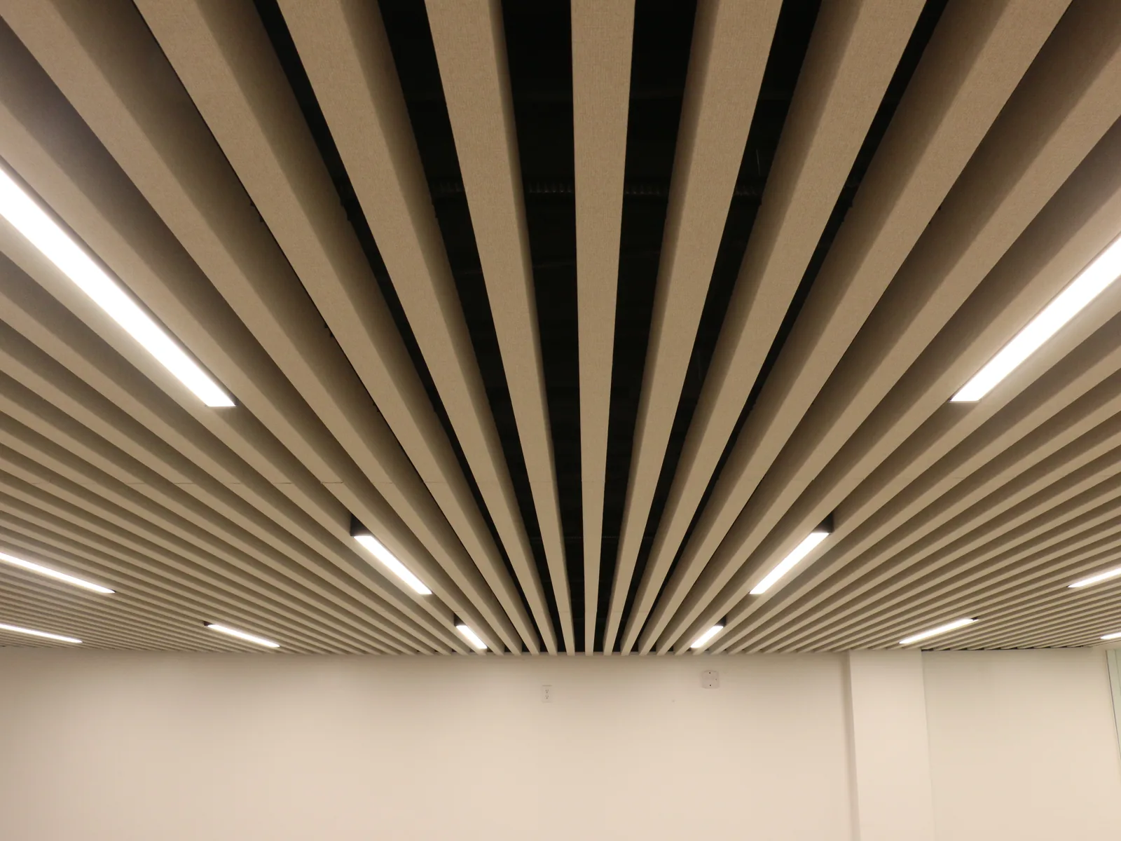 Ceiling of closely spaced beige baffles with linear lights between them, seen in perspective above a plain wall