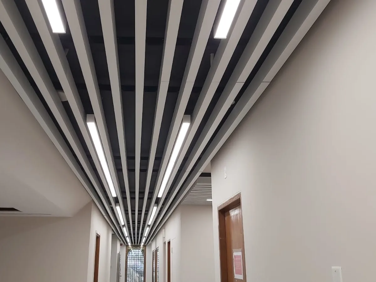 Corridor with a white linear baffle ceiling and strip lights between the blades, ending at a patterned window