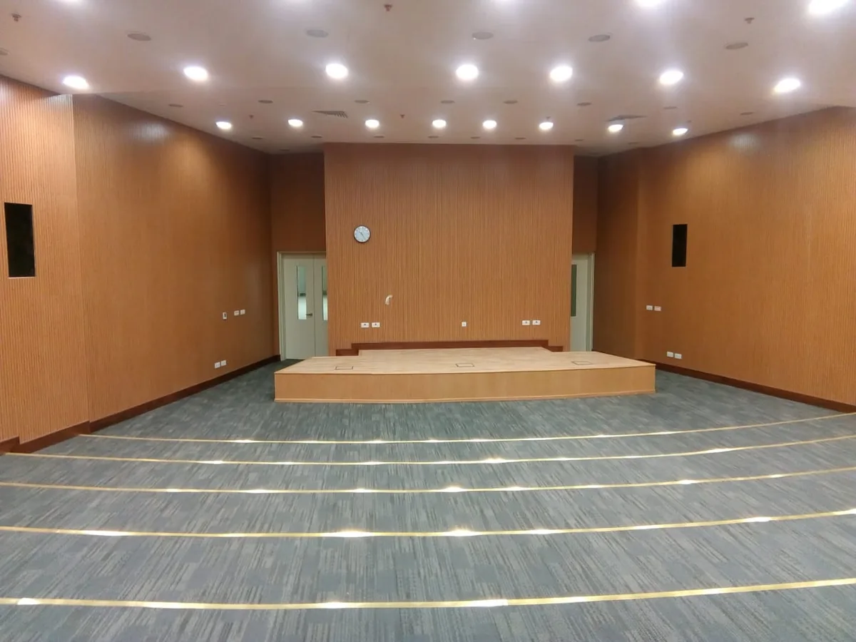 Hall with full-height wood-grain wall panels on three sides, a raised stage and a carpet floor