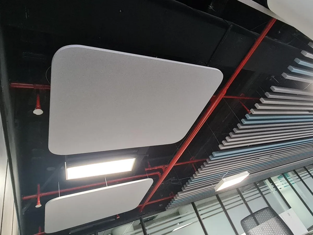 Square acoustic cloud with rounded corners hung below an open soffit with red sprinkler pipes, beside a run of grey and blue baffles