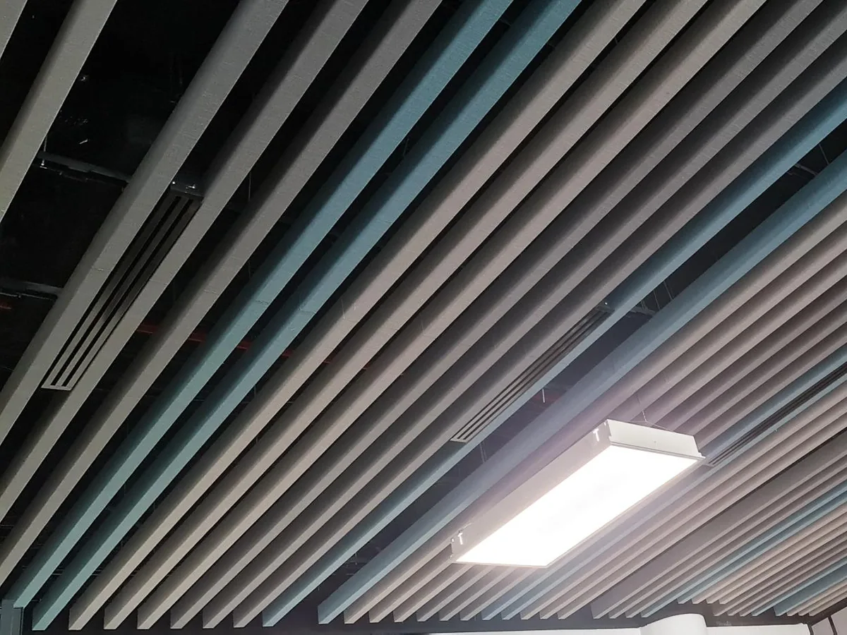 Grey and blue baffles in one run under an open black soffit, with a light fitting and a slot diffuser between the blades