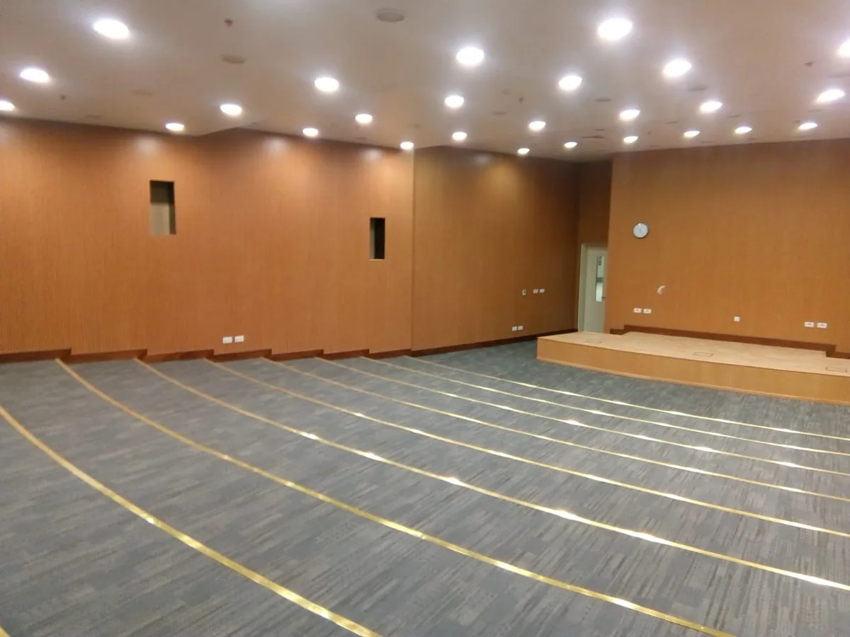 Wide view of a lecture hall: wood-grain wall panels on three sides, a raised stage and curved carpeted tiers with gold-coloured nosings