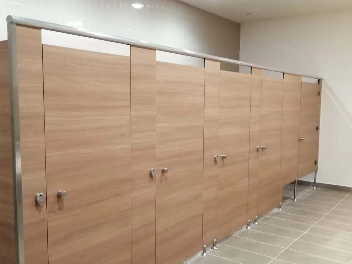 Row of oak-finish toilet cubicle doors braced by a continuous metal headrail, standing on adjustable metal legs