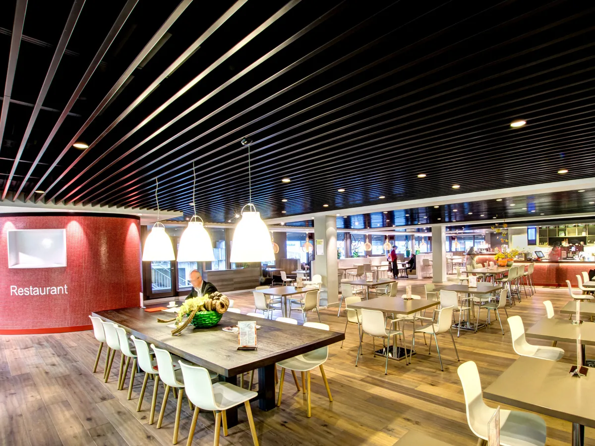 Restaurant dining hall under a dark linear baffle ceiling, with pendant lights over long tables