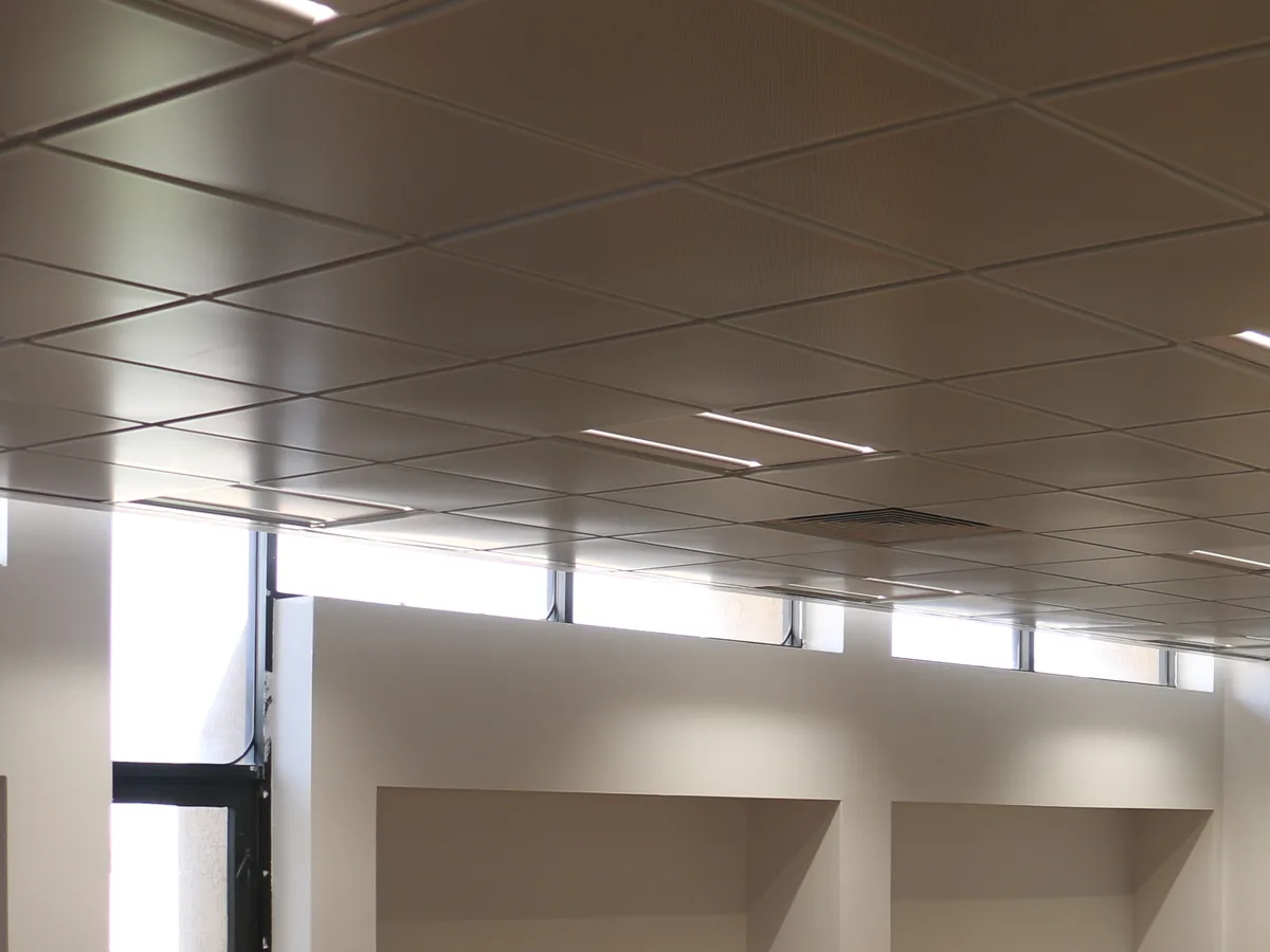Classroom ceiling of lay-in tiles with slim linear lights and an air grille, above a band of high windows