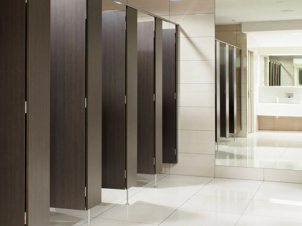 Tall dark wood-grain toilet cubicles on slim metal legs under a headrail, in a bright tiled washroom